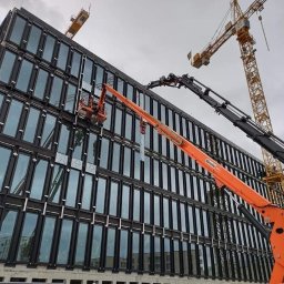 WB-Fassaden - Montaż okien na elewacji nowoczesnego biurowca z użyciem podnośnika koszowego, w tle widoczne dźwigi budowlane i pochmurne niebo.