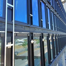 WB-Fassaden - Budynek komercyjny z czarną elewacją i niebieskimi, zabezpieczonymi oknami podczas montażu, widoczne elementy mocujące i rusztowanie.