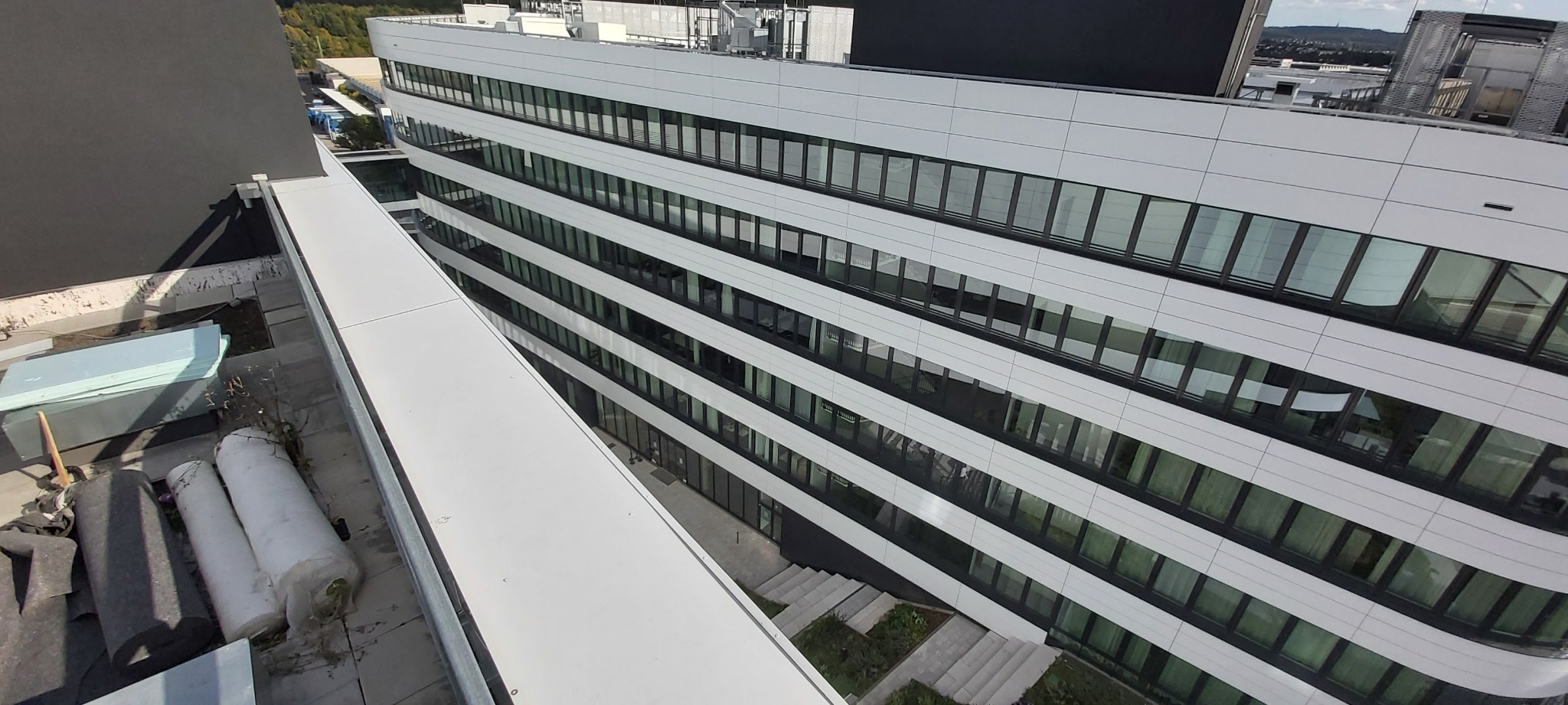 Widok z góry na fragment dachu budynku biurowego z widocznymi materiałami izolacyjnymi w rolkach oraz płytami styropianowymi, z perspektywy dachu sąsiedniego budynku.