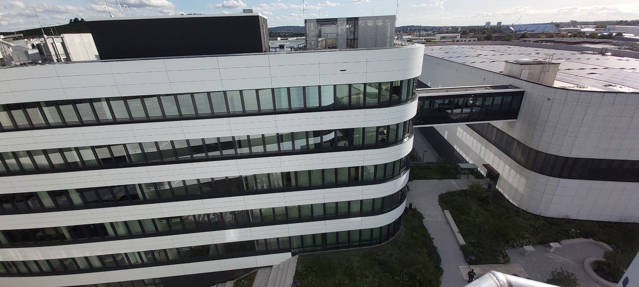 Nowoczesny budynek biurowy z białą elewacją i szklanymi oknami, połączony przeszklonym łącznikiem z halą z panelami słonecznymi na dachu, widok z góry.