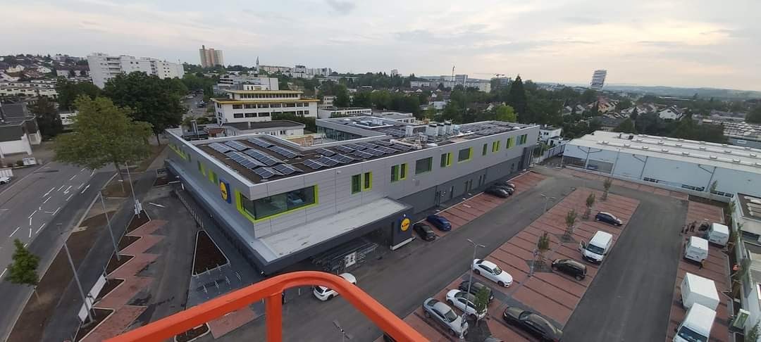 Widok z góry na budynek sklepu z panelami słonecznymi na dachu, parkingiem i otaczającą zabudową miejską w tle.