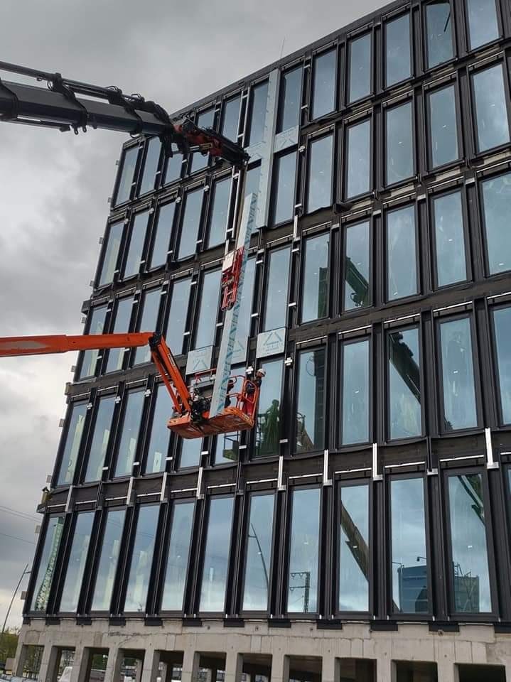 Montaż paneli elewacyjnych na wysokim biurowcu z użyciem podnośnika koszowego i dźwigu, widoczne elementy konstrukcji budynku oraz pracownicy w kaskach.