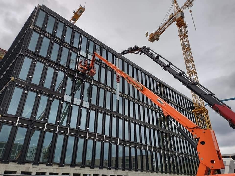 Montaż okien na elewacji nowoczesnego biurowca z użyciem podnośnika koszowego, w tle widoczne dźwigi budowlane i pochmurne niebo.
