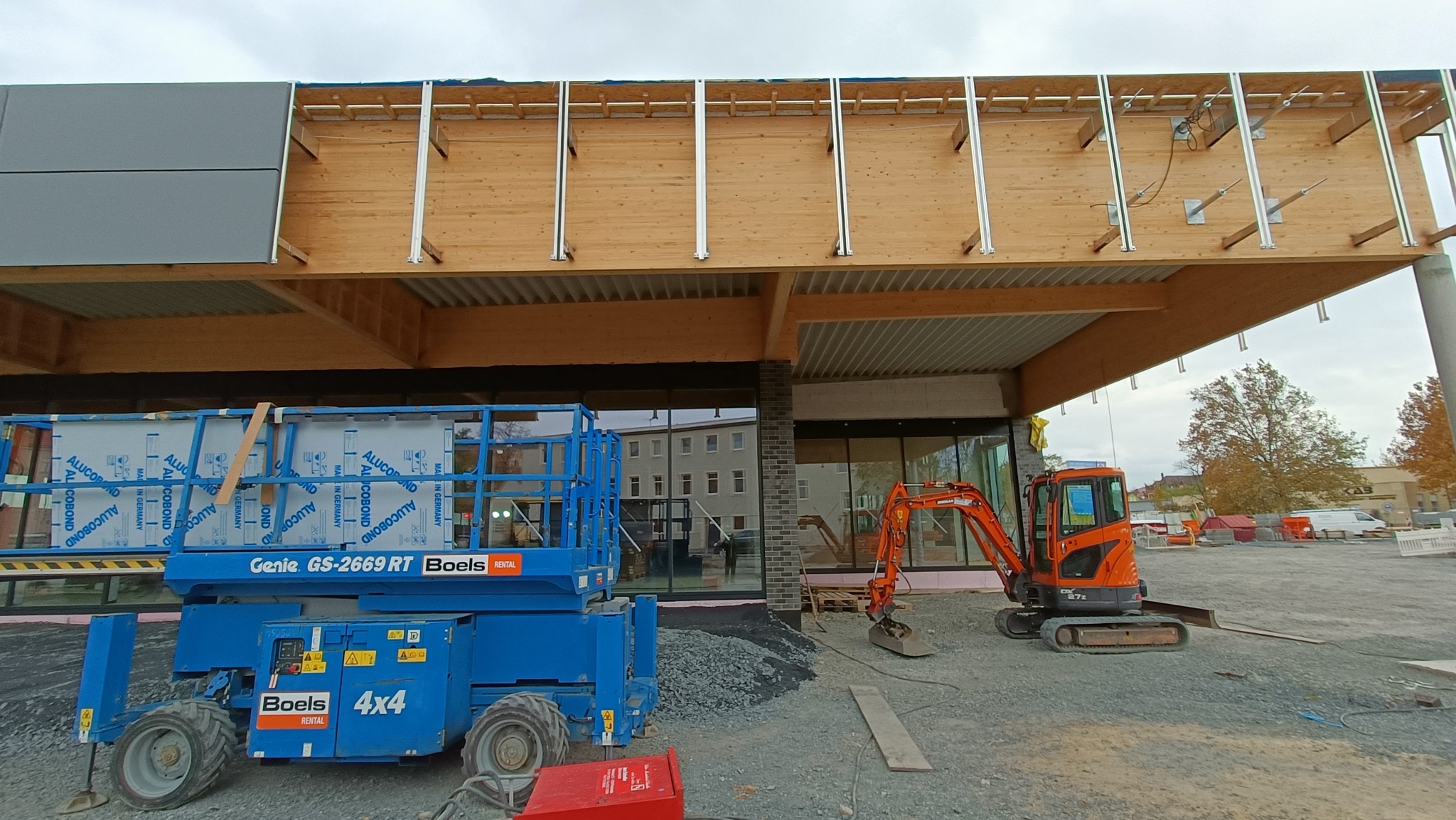 Budowa hali: widok na konstrukcję drewnianą z zamontowanymi elementami stalowymi, z podnośnikiem koszowym Genie i małą koparką Doosan na placu budowy.