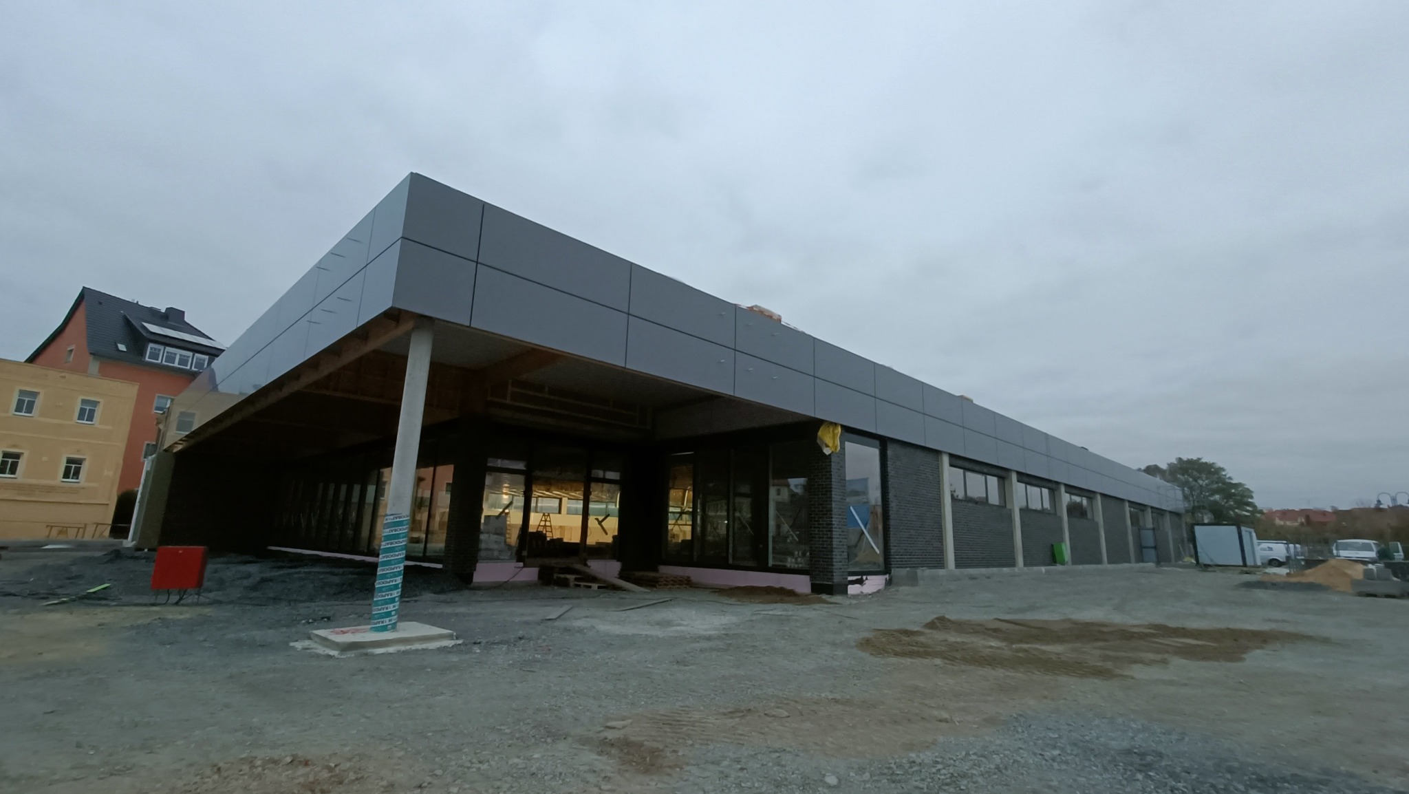 Nowoczesny budynek w trakcie budowy z szarą elewacją z paneli, widoczne elementy konstrukcyjne i rusztowania wewnątrz, teren wokół budynku przygotowany pod zagospodarowanie, pochmurne niebo.