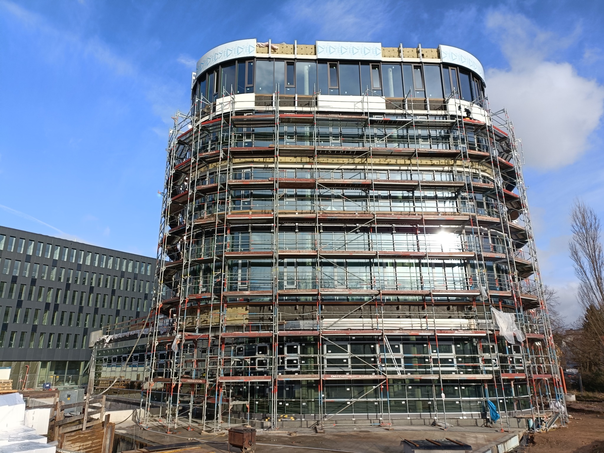 Wielopiętrowy budynek biurowy w trakcie montażu elewacji, otoczony rusztowaniami, widoczny fragment sąsiedniego budynku i błękitne niebo z chmurami.