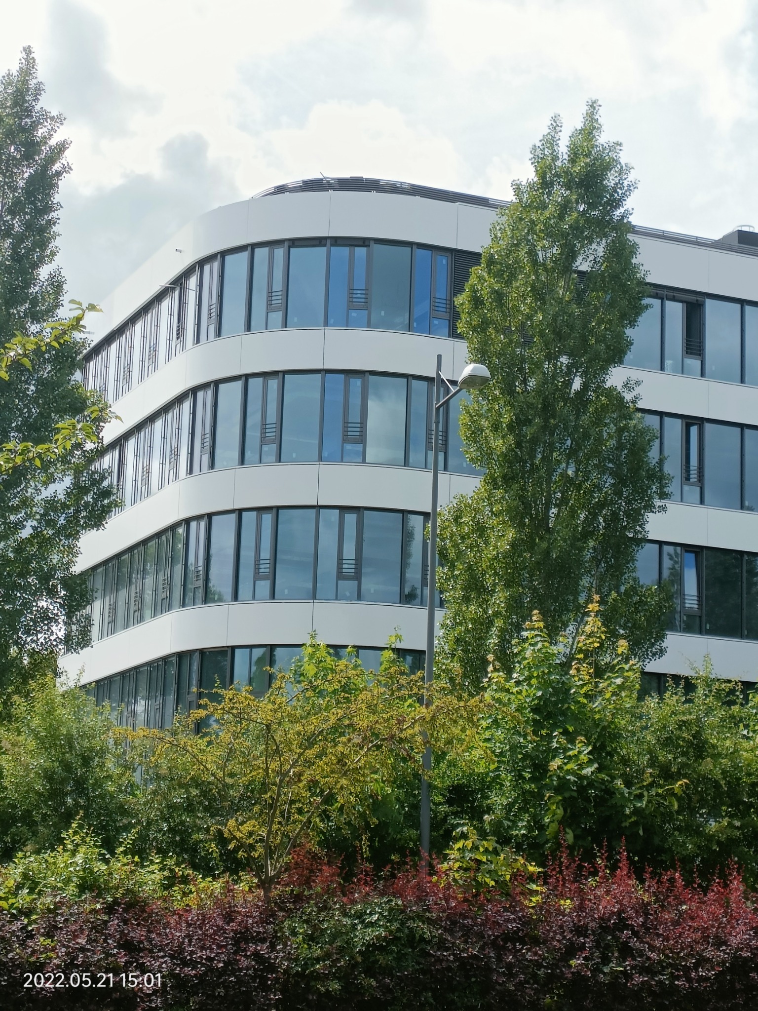 Nowoczesny budynek biurowy z zaokrąglonymi narożnikami i elewacją z paneli, widziany zza zieleni miejskiej, z widoczną lampą uliczną.