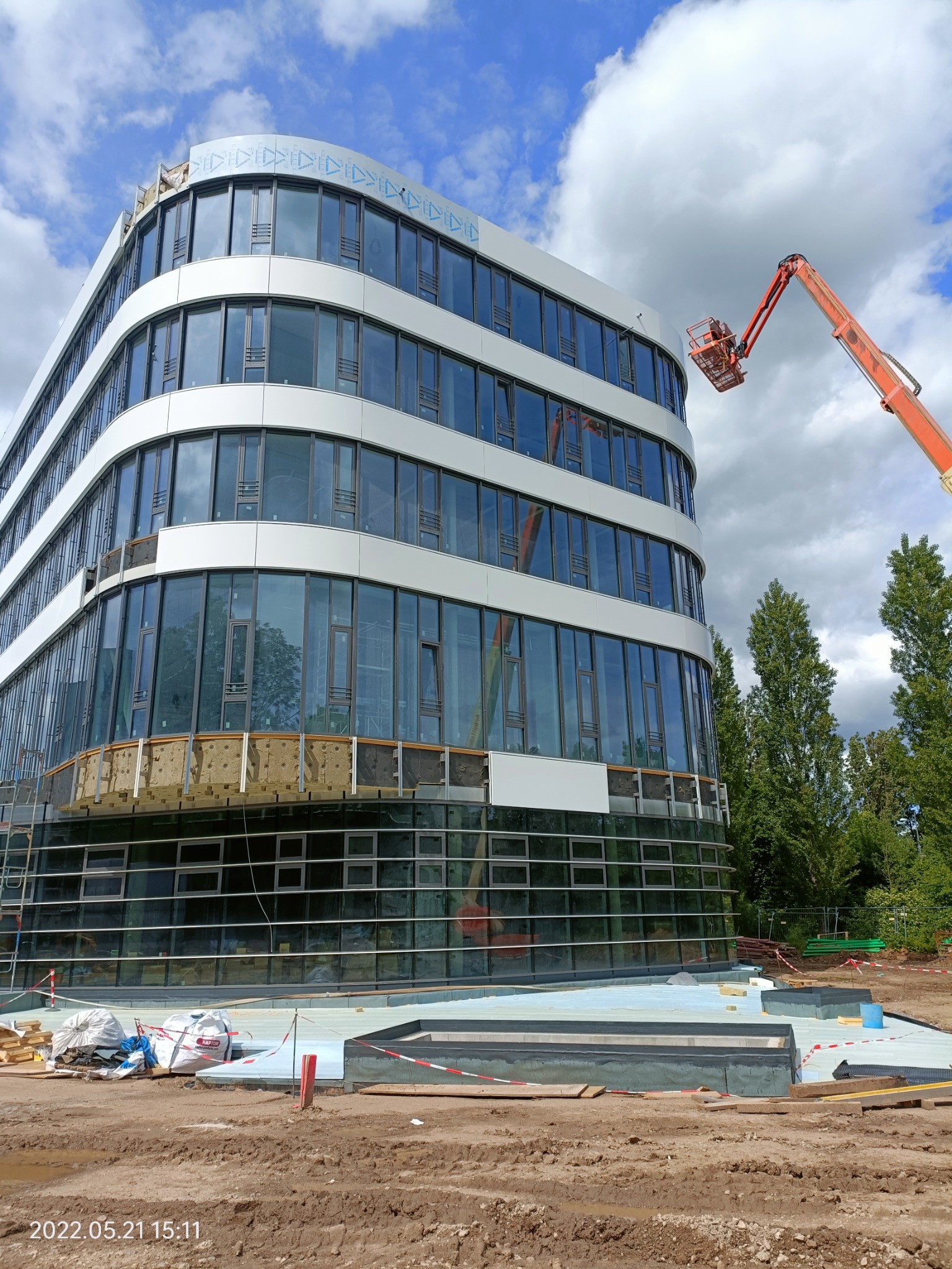 Budynek biurowy w trakcie budowy, widoczne warstwy izolacji na elewacji, podnośnik koszowy przy ścianie, teren budowy z materiałami izolacyjnymi i foliami zabezpieczającymi.
