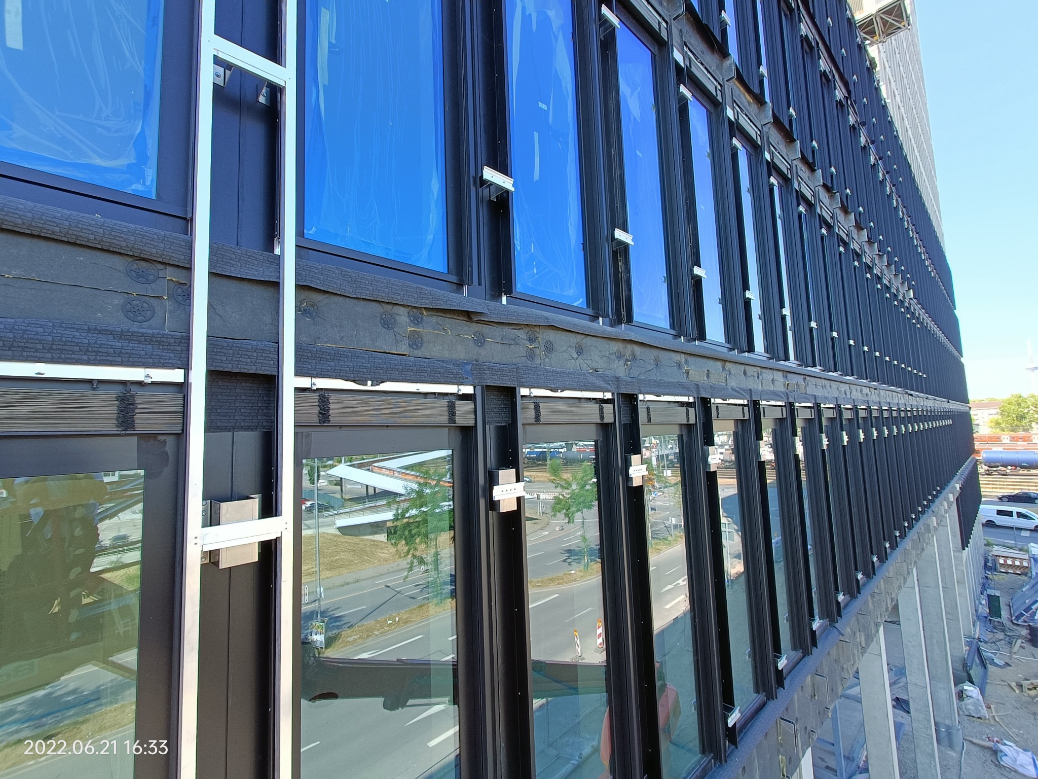 Budynek komercyjny z czarną elewacją i niebieskimi, zabezpieczonymi oknami podczas montażu, widoczne elementy mocujące i rusztowanie.