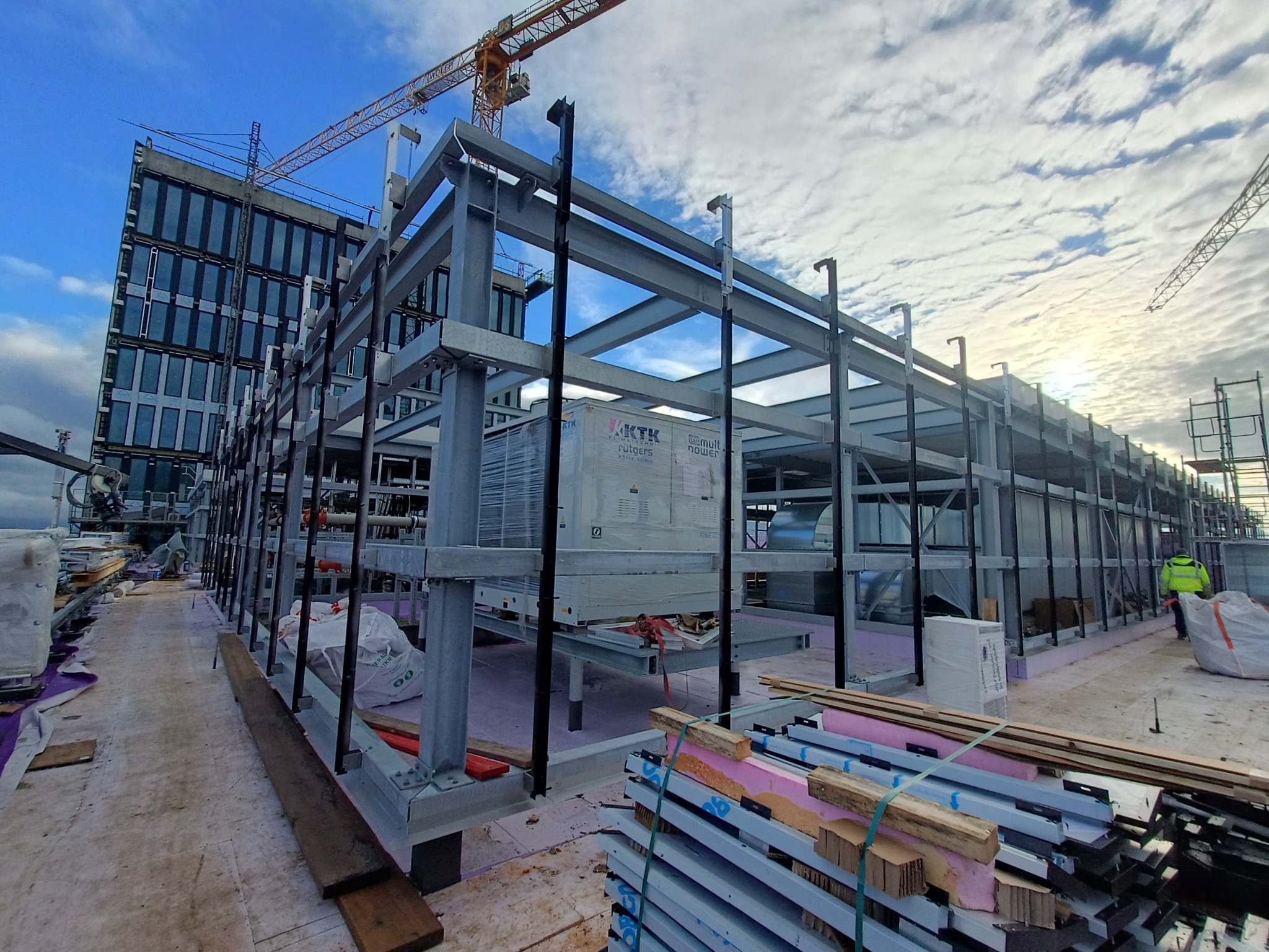 Wznoszenie stalowej konstrukcji hali w Jabłonnie, widoczne elementy wentylacji, dźwig w tle i pracownik w odblaskowej kamizelce.