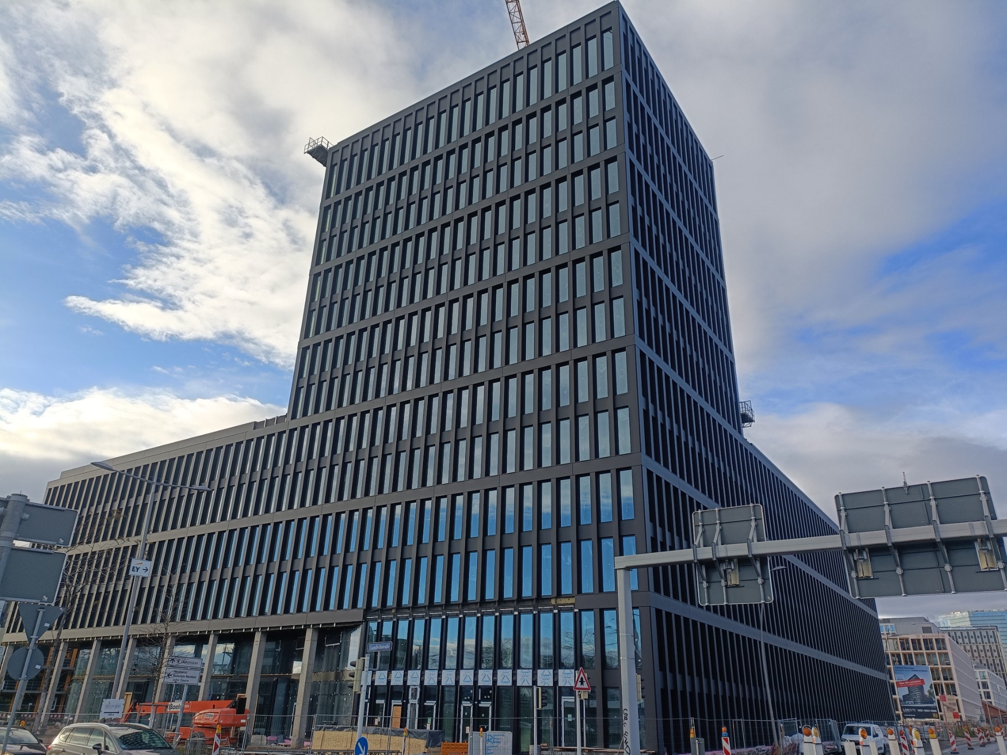 Nowoczesny, wysoki biurowiec z czarną fasadą i licznymi oknami, sfotografowany z poziomu ulicy, z widocznymi znakami drogowymi i elementami placu budowy u dołu.