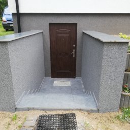 Wykończenia wnętrz i elewacji Marcin Chrzan - Zewnętrzne schody z szarych płytek prowadzące do brązowych drzwi wejściowych w zagłębieniu budynku z szarym, teksturowanym wykończeniem.