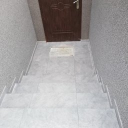 Wykończenia wnętrz i elewacji Marcin Chrzan - Wykończone kafelkami schody prowadzące do brązowych drzwi wejściowych z metalową kratką wentylacyjną w posadzce.