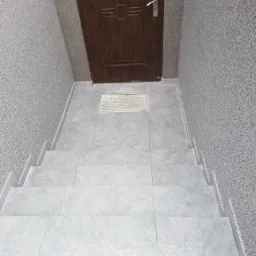 Wykończone kafelkami schody prowadzące do brązowych drzwi wejściowych z metalową kratką wentylacyjną w posadzce.