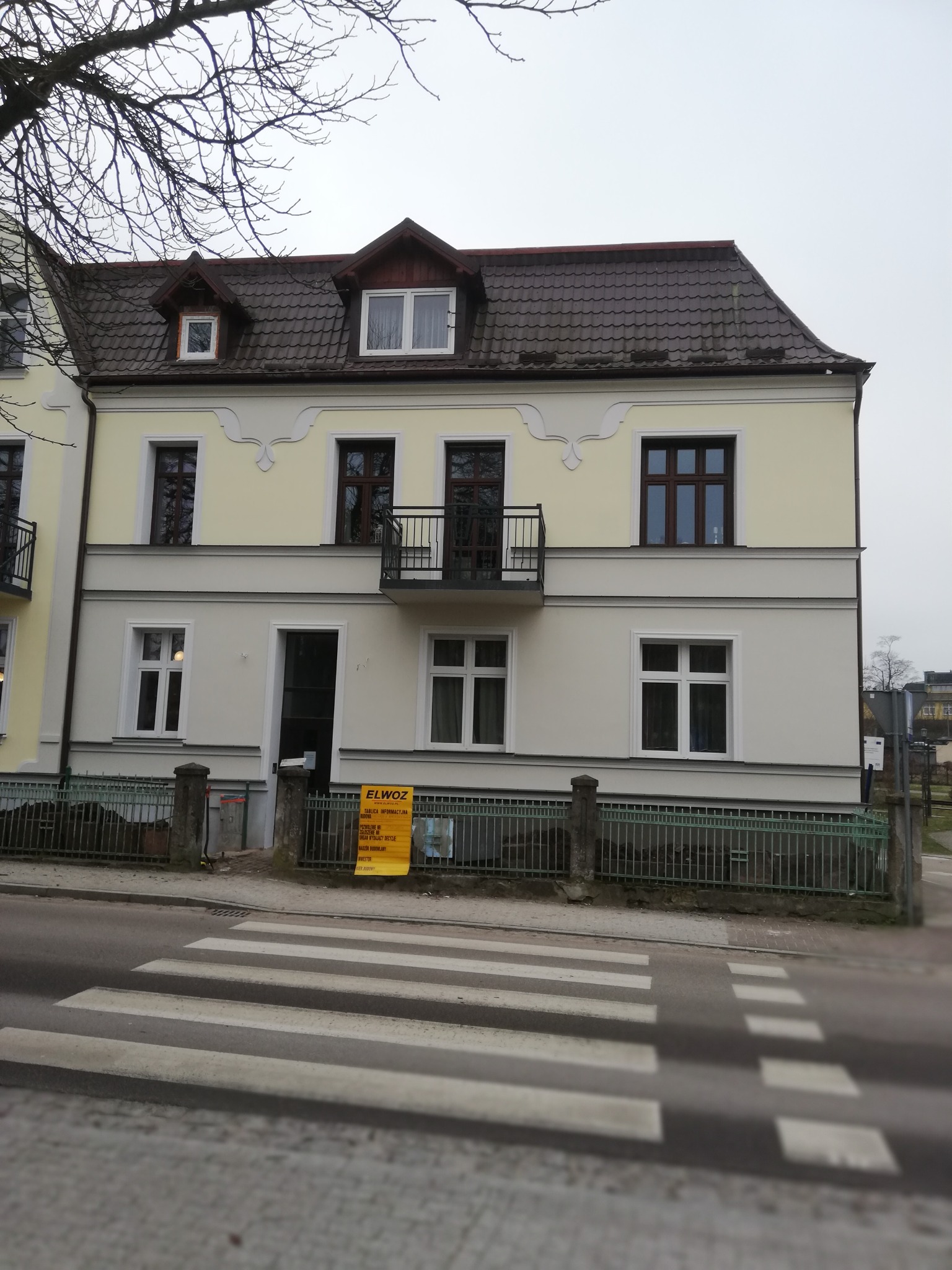 Odnowiona kamienica z żółtą elewacją, brązowymi oknami i balkonem, widziana z poziomu przejścia dla pieszych.