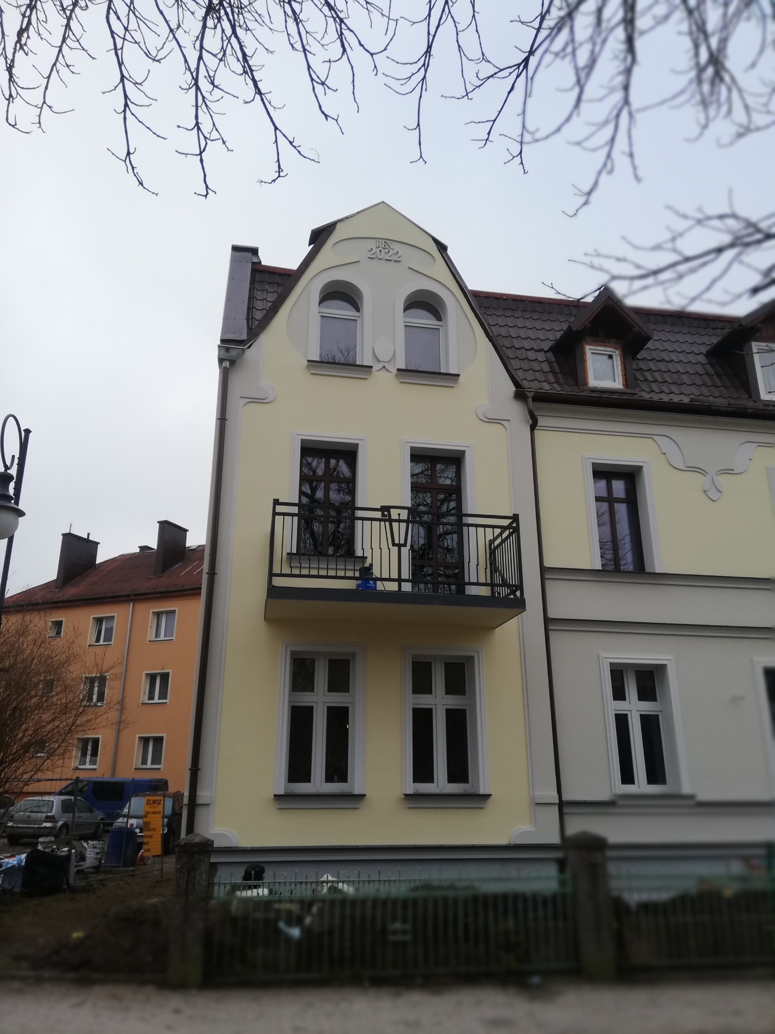 Odnowiona fasada kamienicy w pastelowych kolorach z balkonem i ozdobnymi elementami architektonicznymi, widoczne gałęzie drzew na tle pochmurnego nieba.