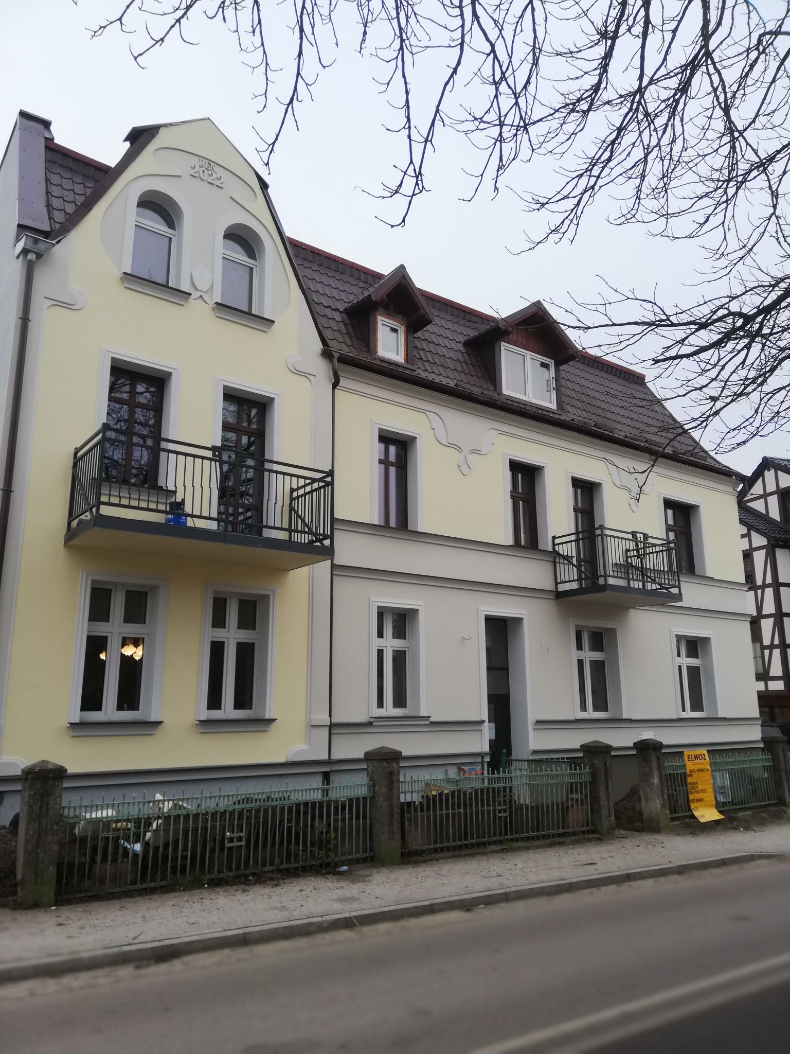 Odnowiona fasada dwukondygnacyjnego domu z balkonami, jasna kolorystyka z elementami dekoracyjnymi, widoczny fragment ulicy i ogrodzenia.