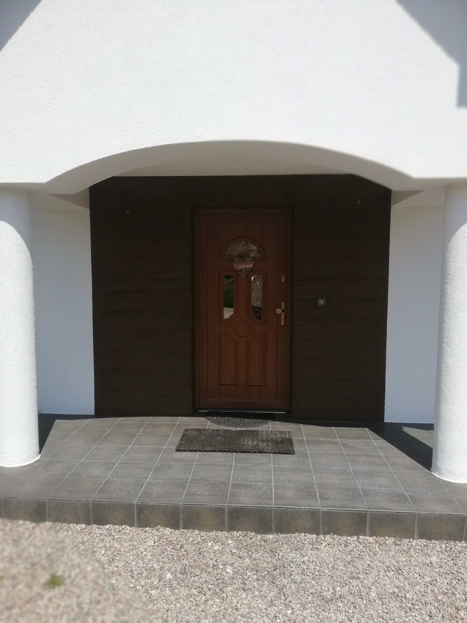 Wejście do domu z brązowymi drzwiami z dekoracyjnym okienkiem 'HOME', ciemną elewacją i białymi kolumnami oraz szarą, wykafelkowaną werandą i żwirowym podłożem.