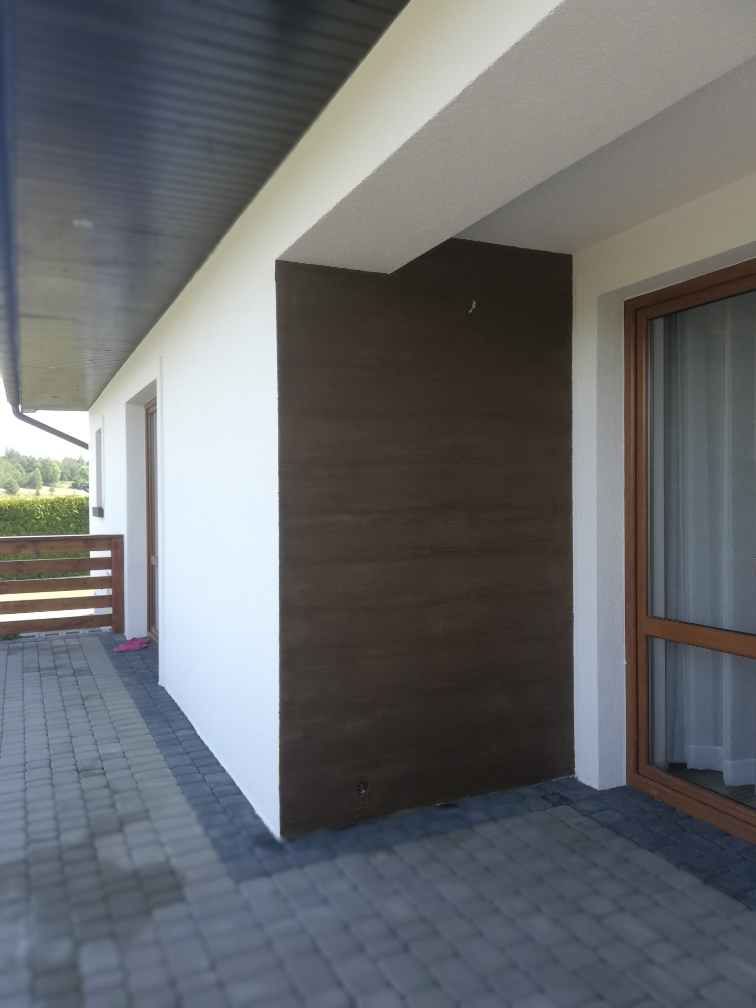 Wykończona elewacja domu z brązową ścianą imitującą drewno, białe ściany, okno z brązową ramą i szara kostka brukowa na tarasie.