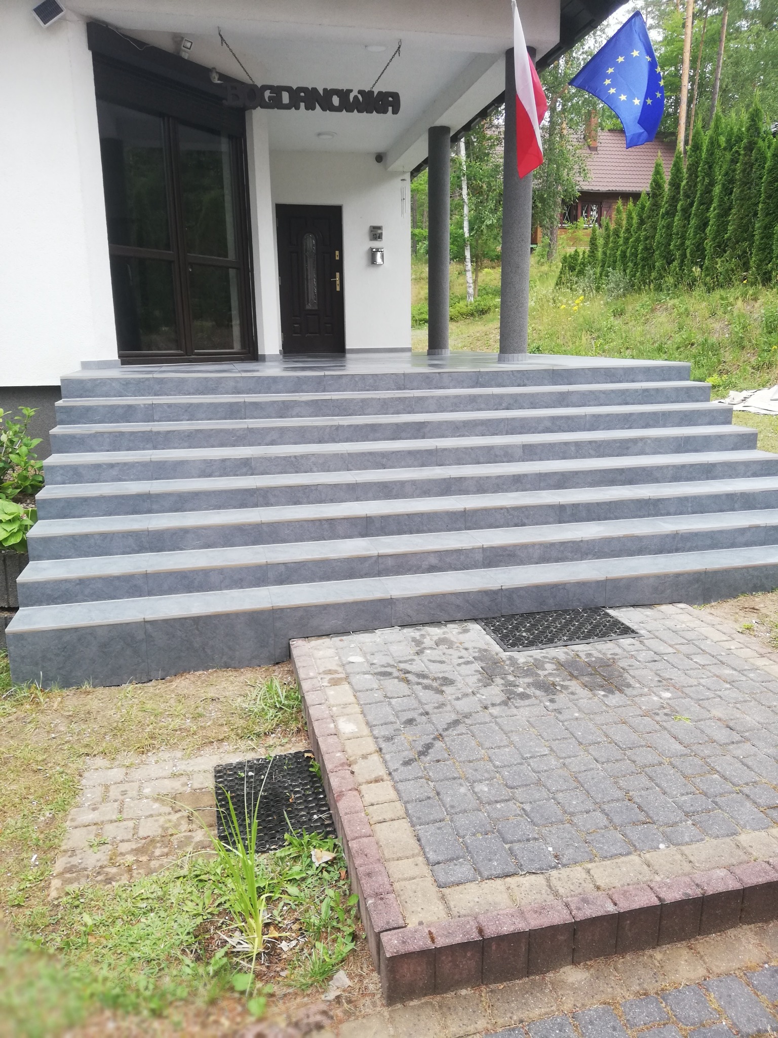 Zewnętrzne schody wejściowe do budynku wyłożone szarymi płytkami, prowadzące do drzwi wejściowych i okna, z widocznymi flagami Polski i Unii Europejskiej.