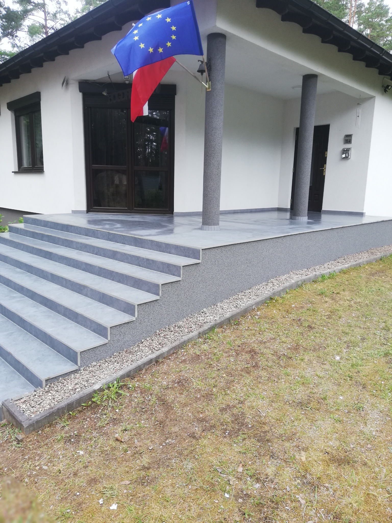 Wykończone wejście do domu z szarymi schodami z płytek, kolumnami i elewacją w odcieniach bieli, udekorowane flagami Unii Europejskiej i Polski.
