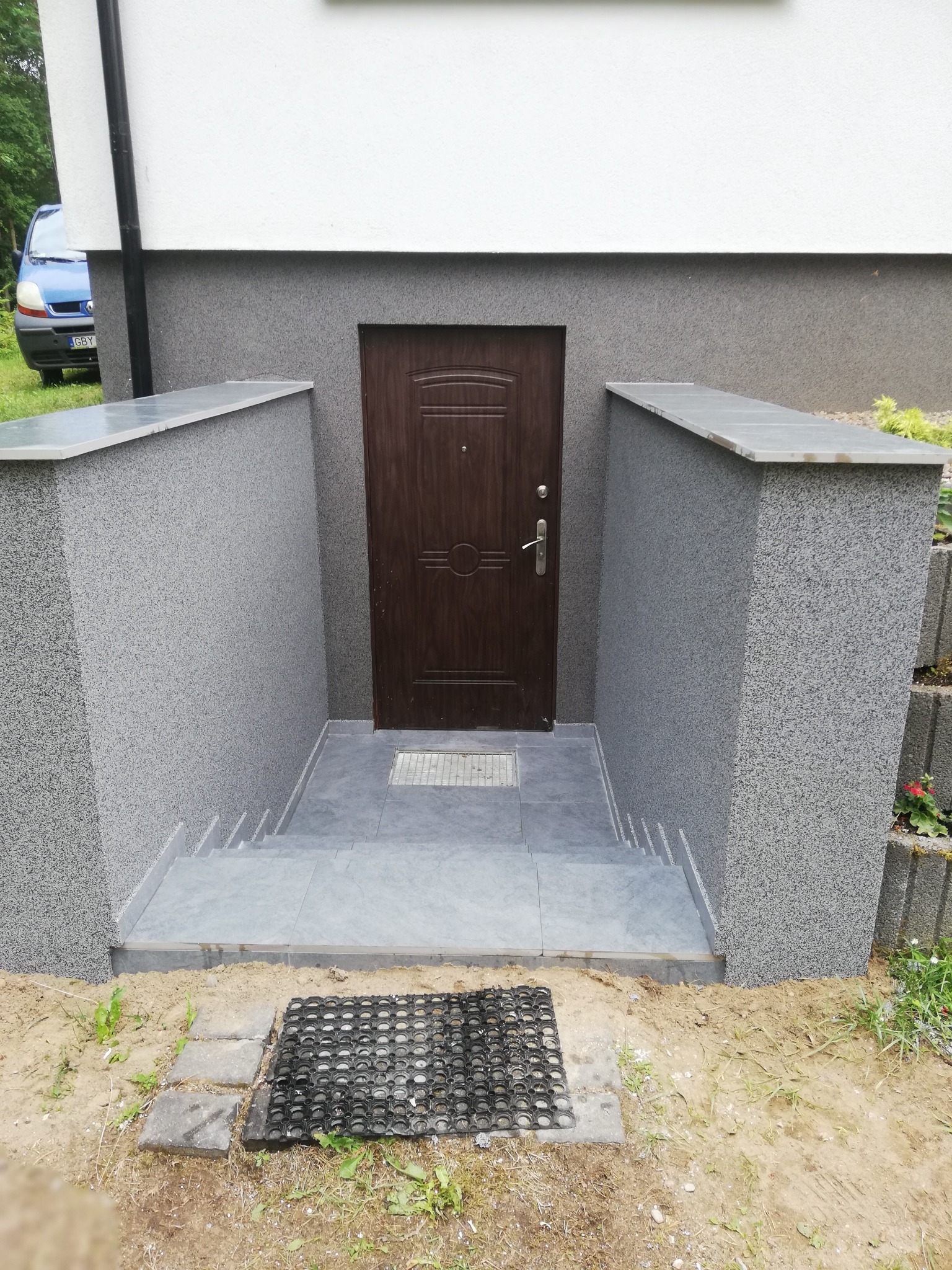 Zewnętrzne schody z szarych płytek prowadzące do brązowych drzwi wejściowych w zagłębieniu budynku z szarym, teksturowanym wykończeniem.