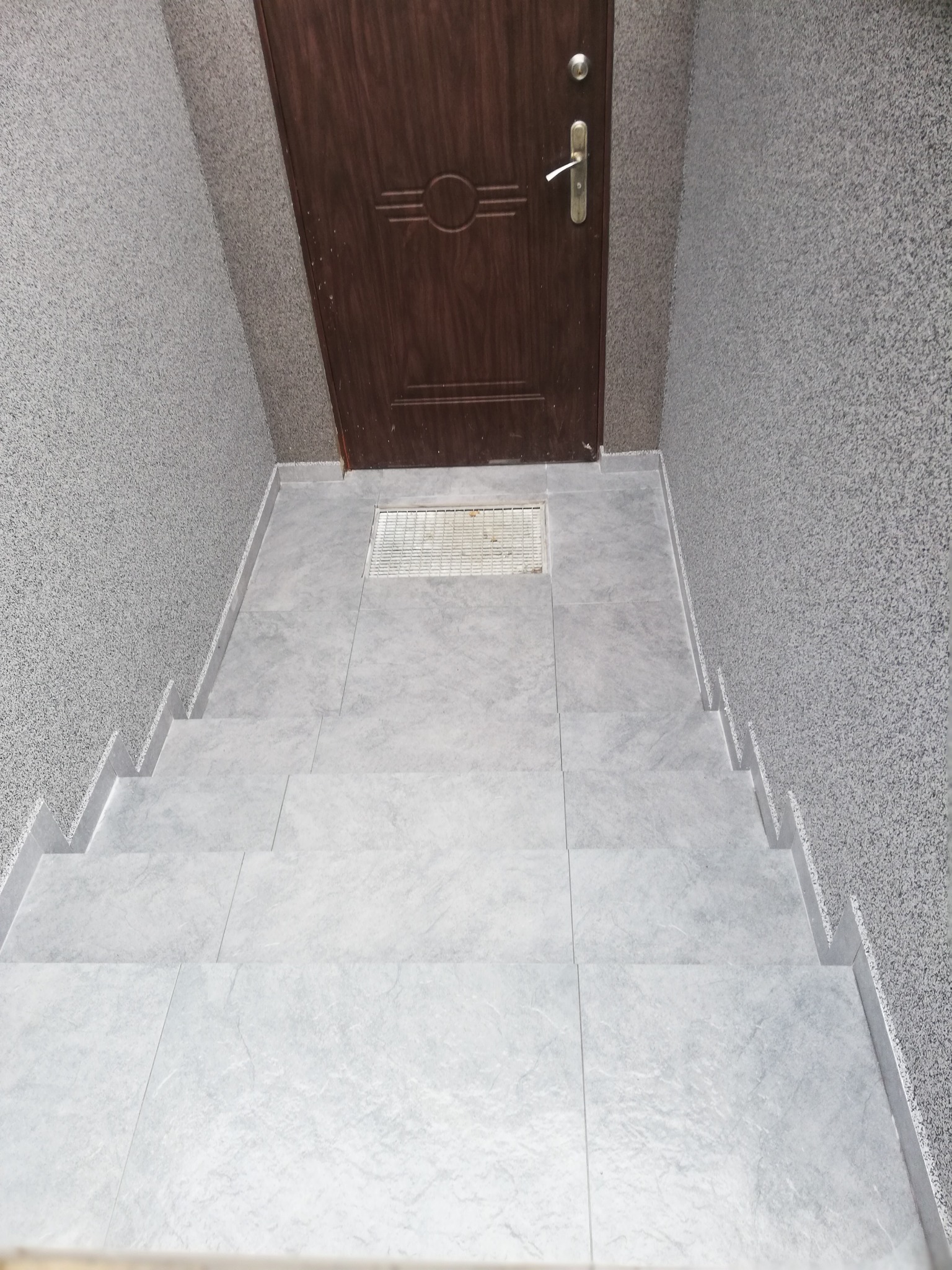 Wykończone kafelkami schody prowadzące do brązowych drzwi wejściowych z metalową kratką wentylacyjną w posadzce.