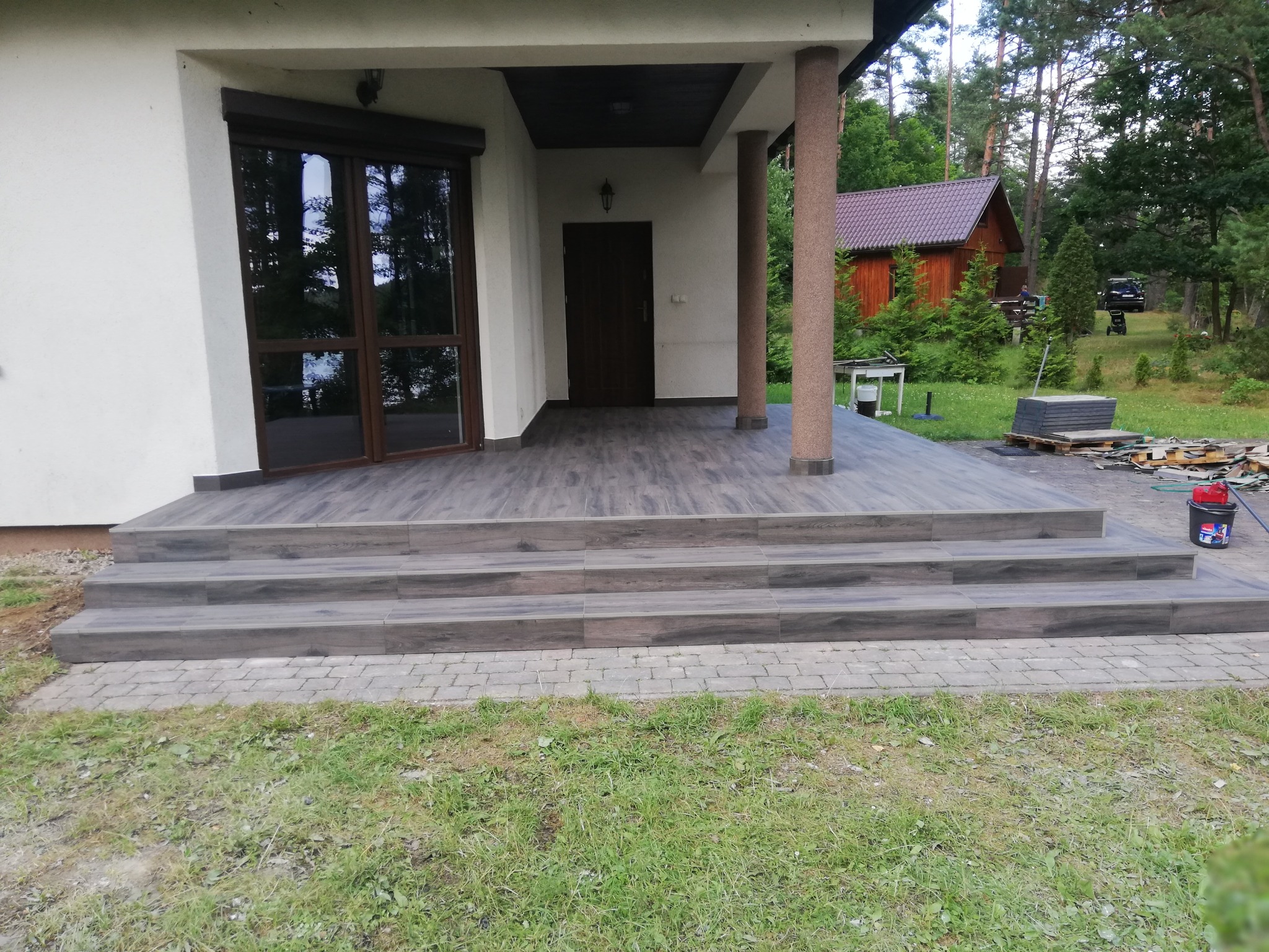 Zadaszone wejście do domu z nowymi schodami wyłożonymi płytkami imitującymi drewno, widoczne resztki materiałów budowlanych i narzędzia.