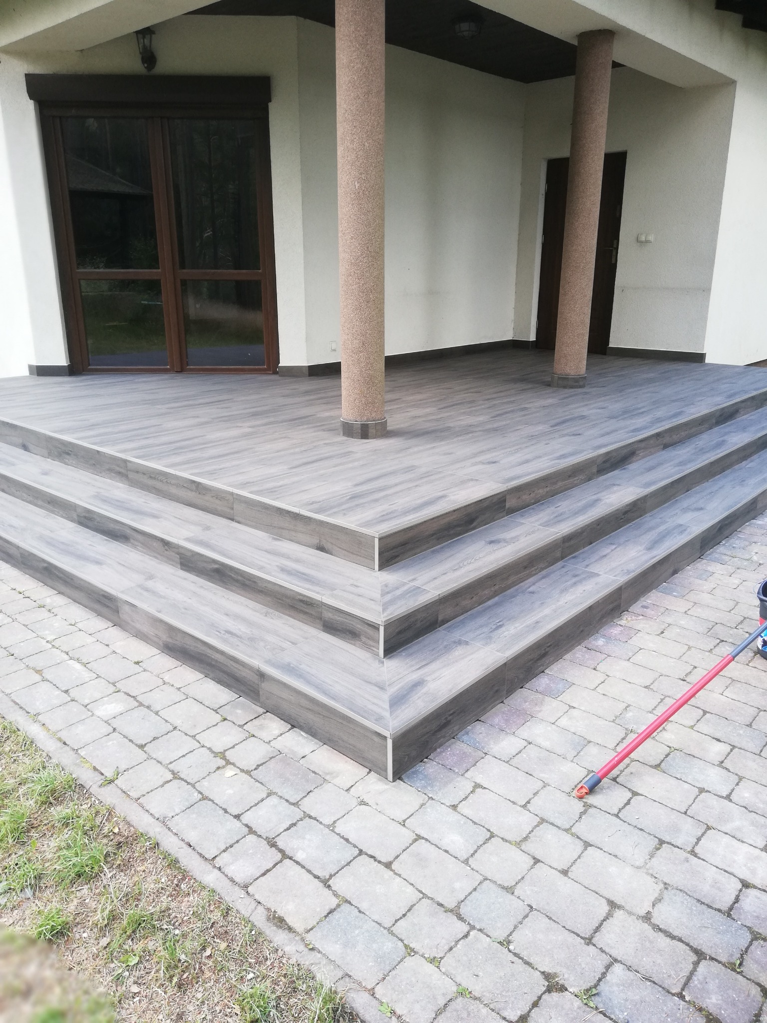 Zewnętrzne schody wejściowe wyłożone szarą płytką imitującą drewno, prowadzące do domu z kolumnami, widok z poziomu kostki brukowej.