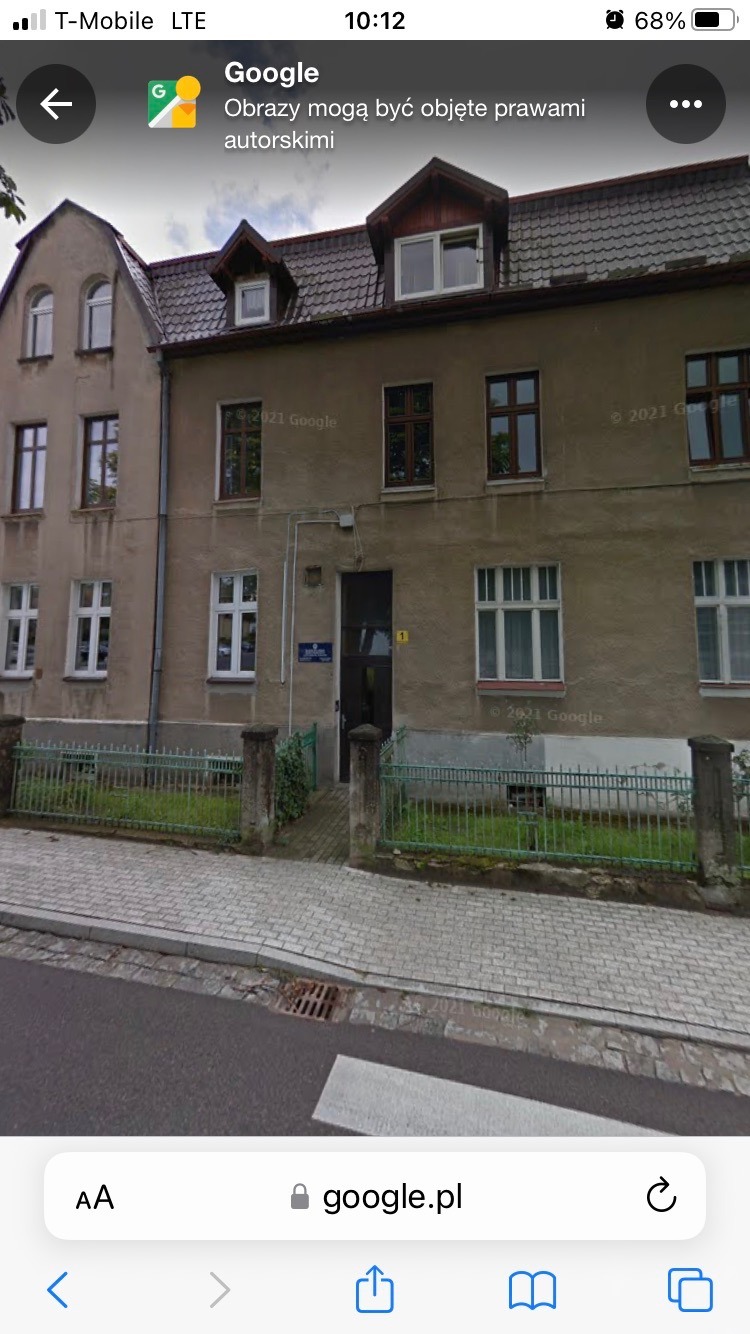 Fasada dwupiętrowej kamienicy z licznymi oknami, ciemnymi drzwiami wejściowymi z numerem 1 i zielonym ogrodzeniem od ulicy.