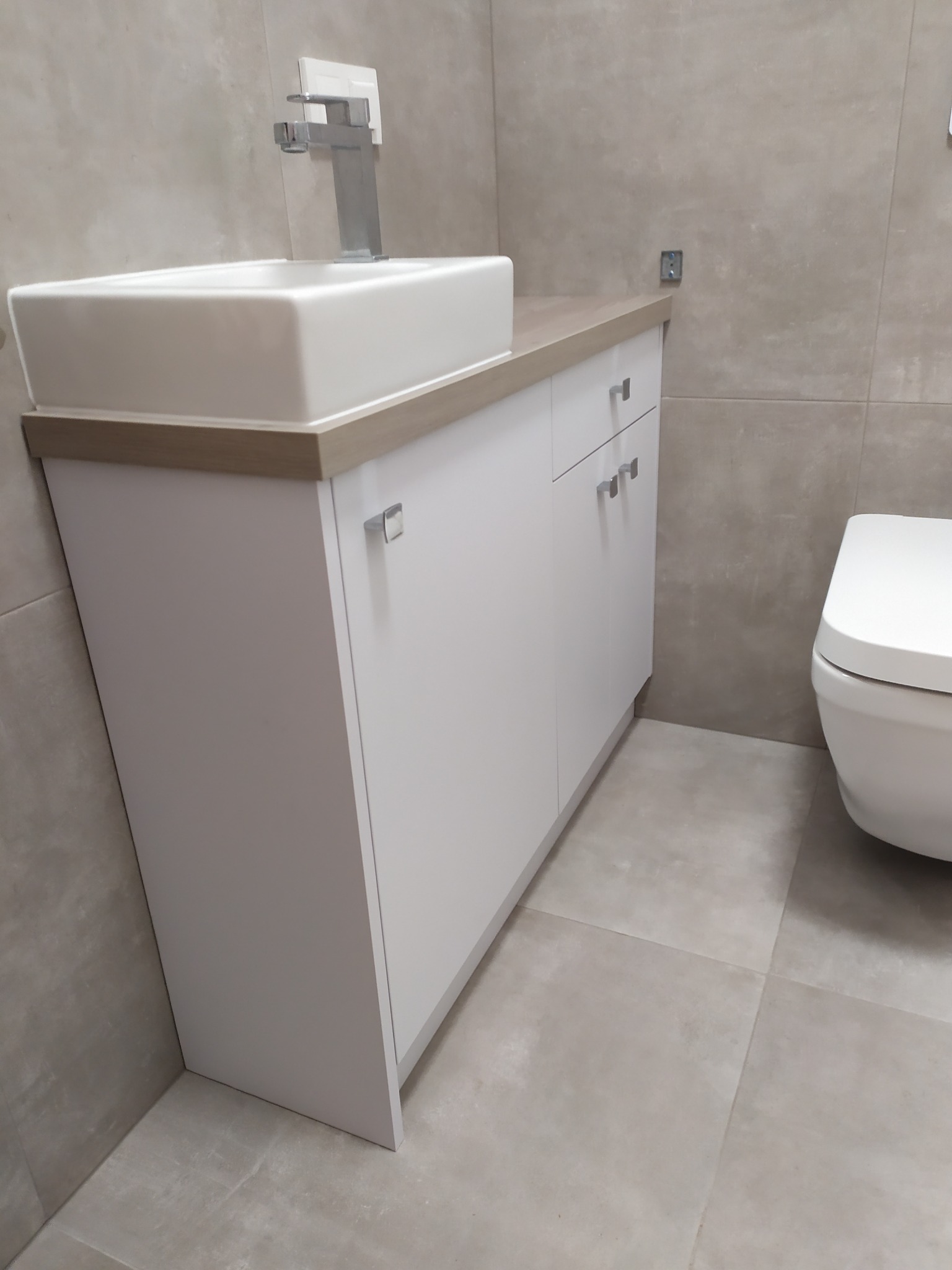 Biała szafka pod umywalkę nablatową z drewnianym blatem w nowoczesnej łazience z szarymi płytkami.