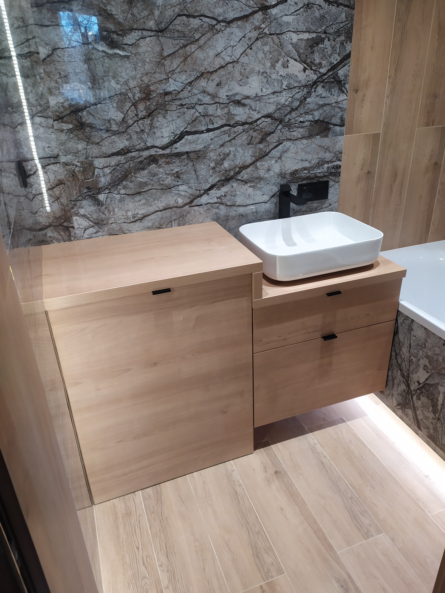 Nowoczesna łazienka z jasnym drewnianym blatem, nablatową białą umywalką i czarną armaturą na tle ściany z imitacji marmuru.