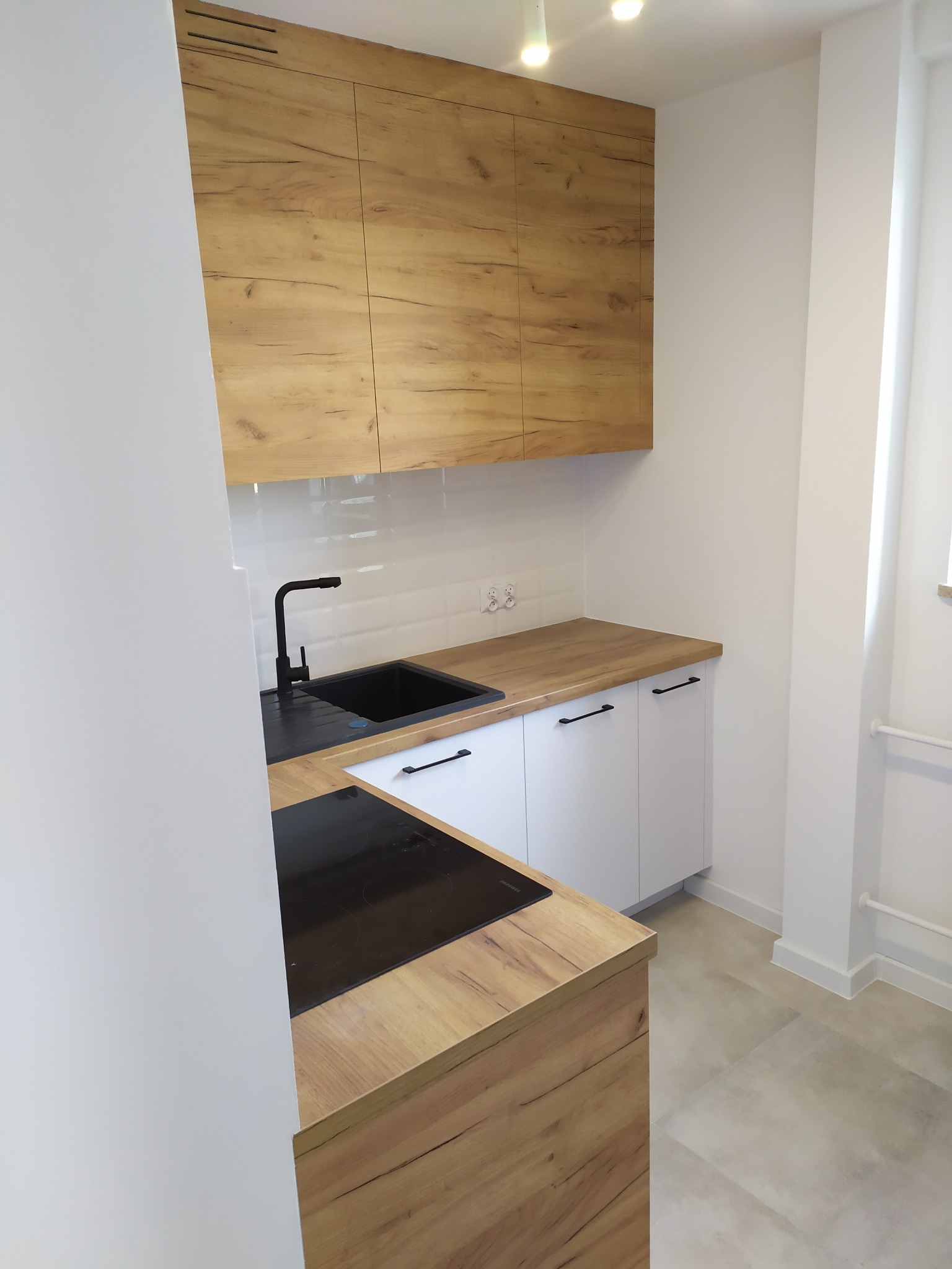 Nowoczesna, jasna kuchnia z drewnianymi szafkami górnymi i blatem w kolorze drewna, czarnym zlewem i płytą indukcyjną, zrealizowana w Nowym Dworze Mazowieckim.