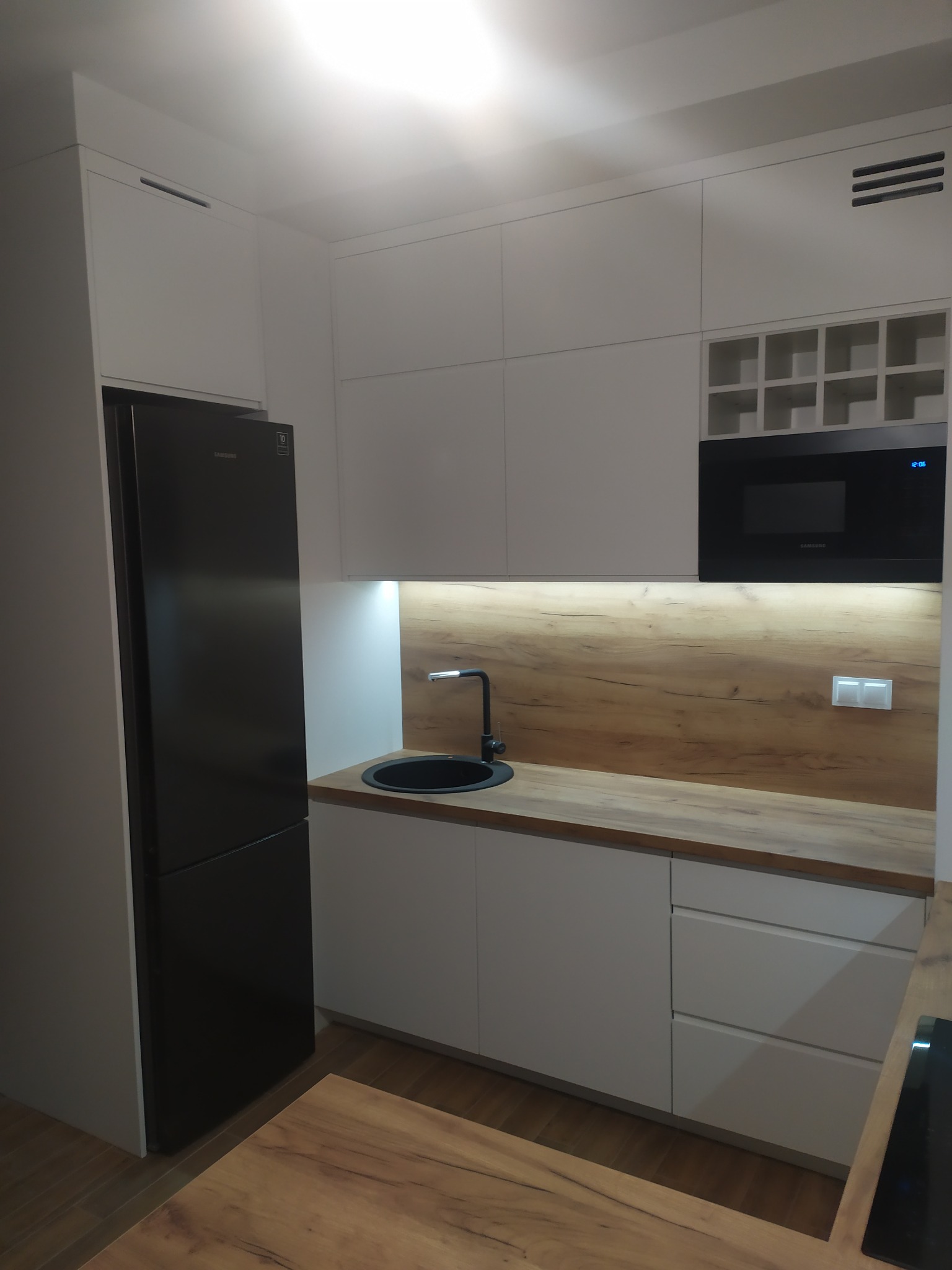 Nowoczesna kuchnia z białymi szafkami, blatem drewnopodobnym, czarnym zlewem i lodówką Samsung, oświetleniem LED pod szafkami wiszącymi i wbudowaną mikrofalówką.