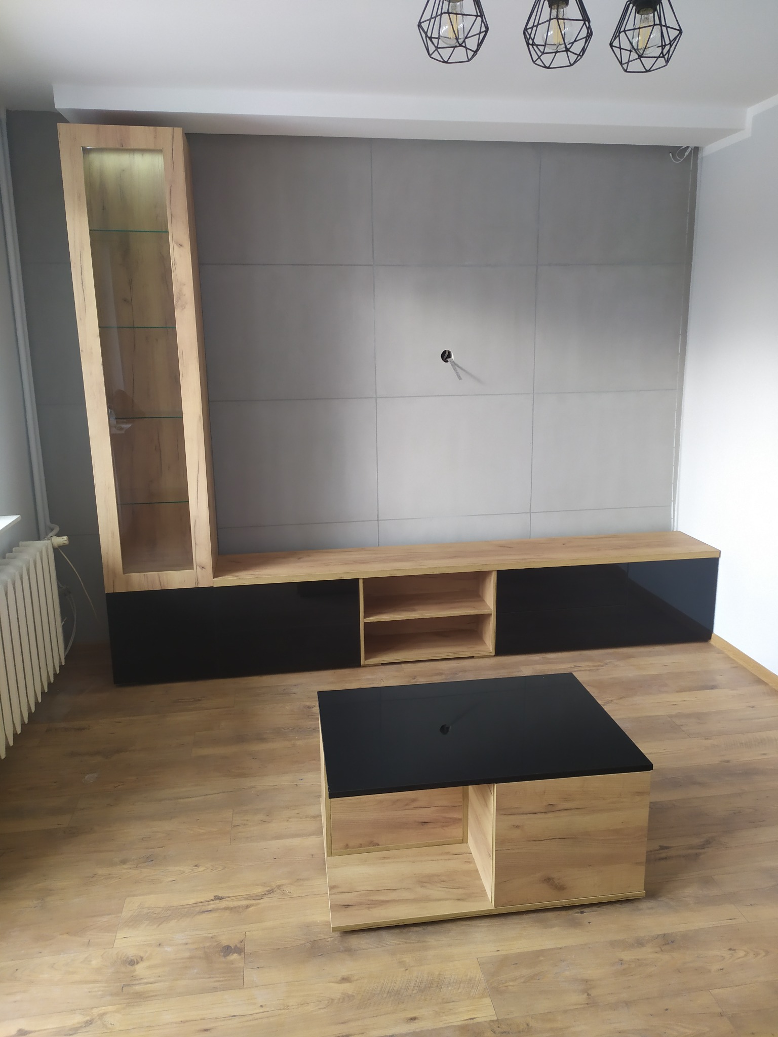 Nowoczesny salon z drewnianą podłogą, meblościanką z elementami czerni i drewna oraz stolikiem kawowym z czarnym blatem, oświetlony trzema lampami w stylu industrialnym.