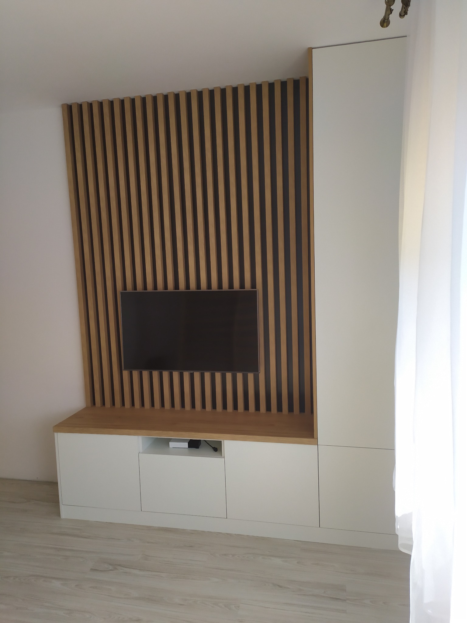 Nowoczesna zabudowa ściany telewizyjnej z drewnianymi lamelami i białymi szafkami, minimalistyczny design.