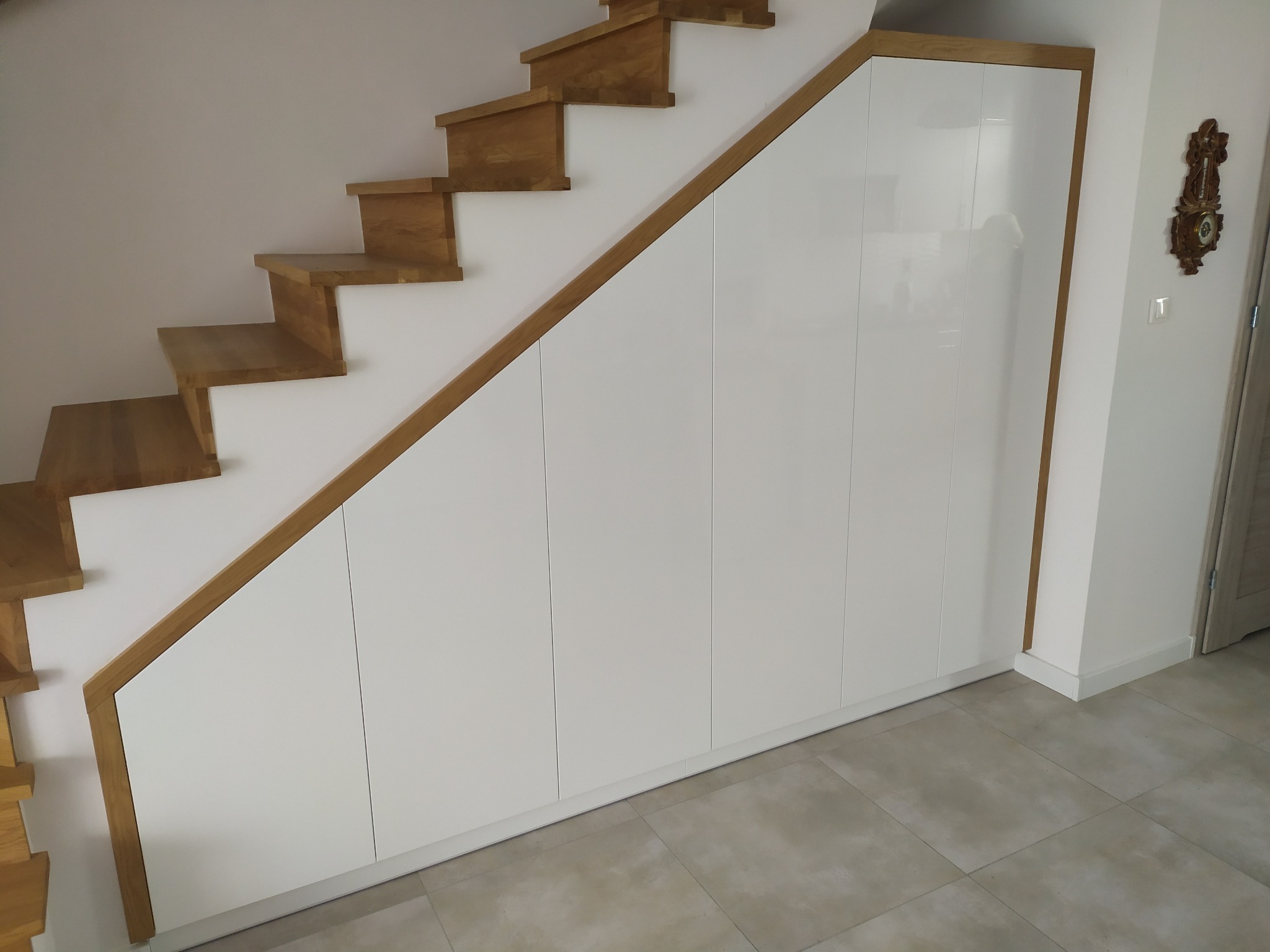 Biała zabudowa pod schodami z drewnianym wykończeniem, widoczne drewniane schody i zegar na ścianie