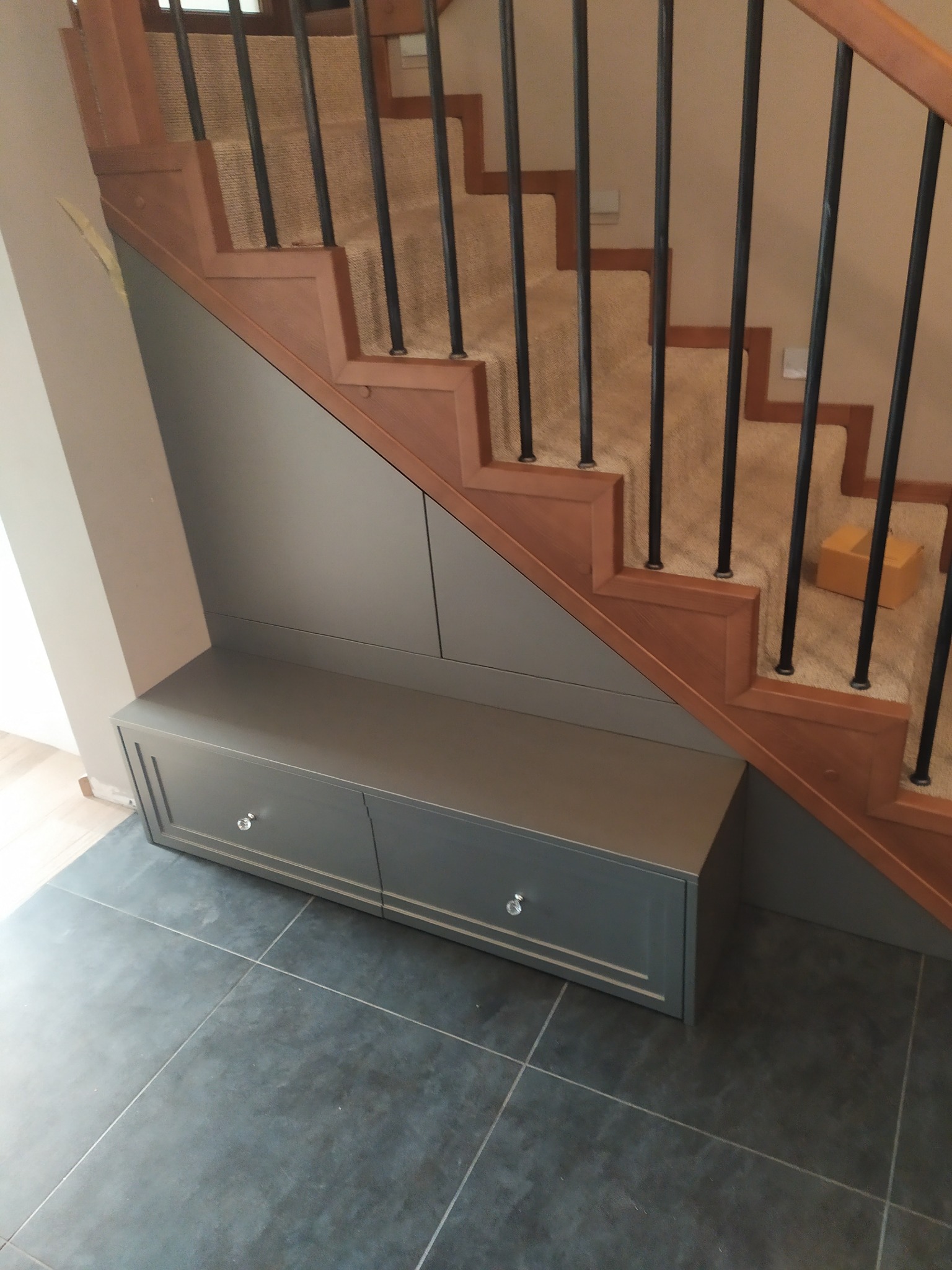Szara zabudowa pod schodami z dwoma szufladami z kryształowymi gałkami, dopasowana kolorystycznie do ściany, na tle drewnianych schodów z czarnymi balustradami i szarą podłogą z płytek.