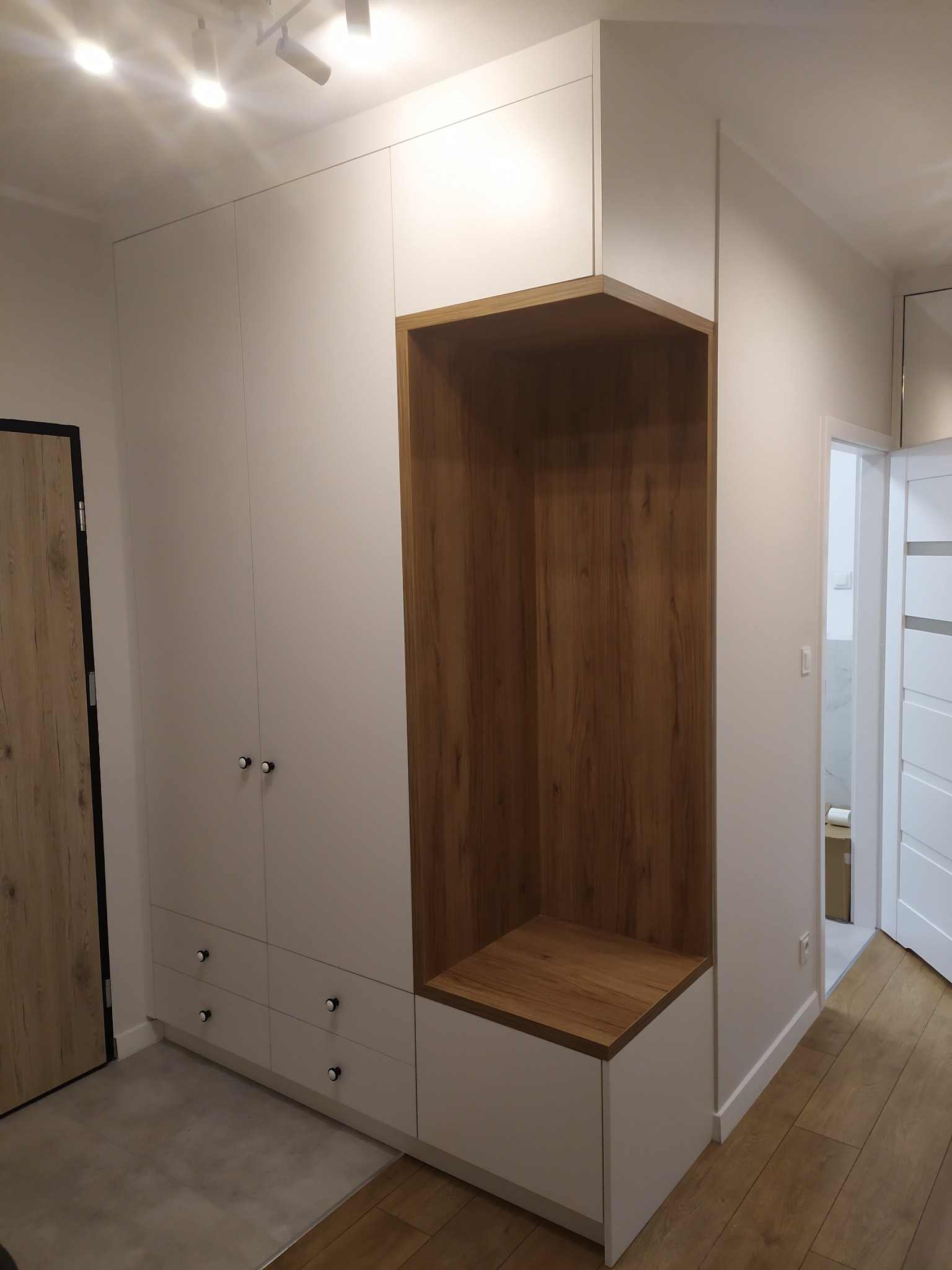 Biała szafa wnękowa z drewnianą wnęką na buty i siedziskiem, zlokalizowana w przedpokoju z szarą podłogą i drewnianą podłogą w dalszej części.