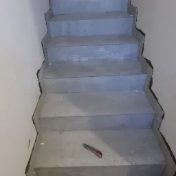 Betonowe schody w trakcie wykańczania, widoczny nóż tapicerski i plastikowe krzyżyki dystansowe na dolnym stopniu.