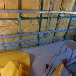 Ocieplenie poddasza wełną mineralną, widoczne profile metalowe, instalacja wodna, płyty gipsowo-kartonowe i narzędzia na podłodze. Fragment okna dachowego w tle.