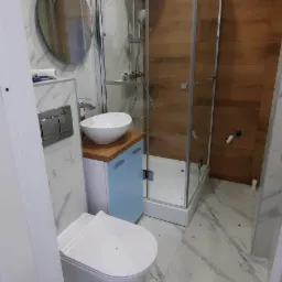 Nowoczesna łazienka z kabiną prysznicową, umywalką nablatową na szafce w kolorze baby blue i podwieszaną toaletą, wykończona płytkami imitującymi marmur i drewno.