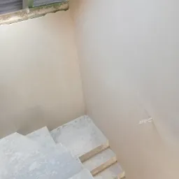 Wnętrze domu w trakcie wykańczania: gładkie, jasne ściany po nałożeniu tynku maszynowego oraz surowe, betonowe schody prowadzące w górę.