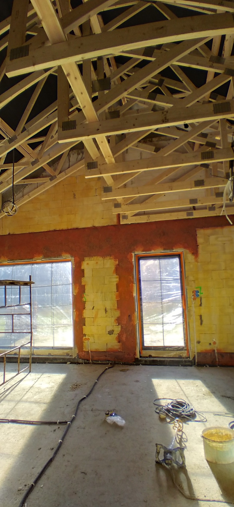 Wnętrze budynku w trakcie prac tynkarskich: ściany z częściowo nałożonym tynkiem w kolorze ceglastym, widoczne okna zabezpieczone folią, drewniana konstrukcja dachu, kable elektryczne i narzędzia...