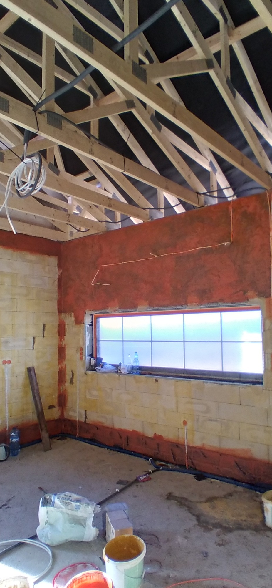 Wnętrze w trakcie prac wykończeniowych: ściana częściowo pokryta czerwoną warstwą, widoczne drewniane elementy konstrukcji dachu, okno z matową szybą, worki z zaprawą i wiaderka na podłodze.