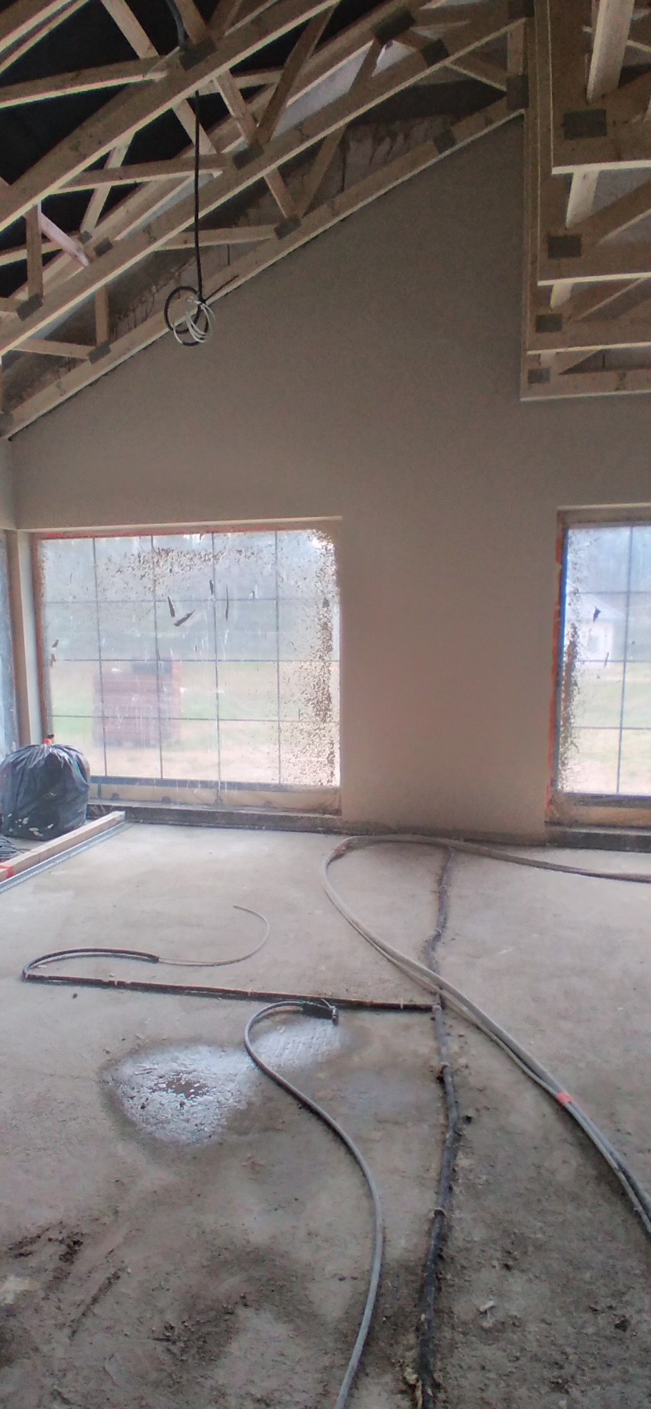 Wnętrze w trakcie prac wykończeniowych, ściany pokryte świeżym tynkiem, widoczne drewniane elementy konstrukcji dachu, zanieczyszczone okna i węże instalacyjne na podłodze.