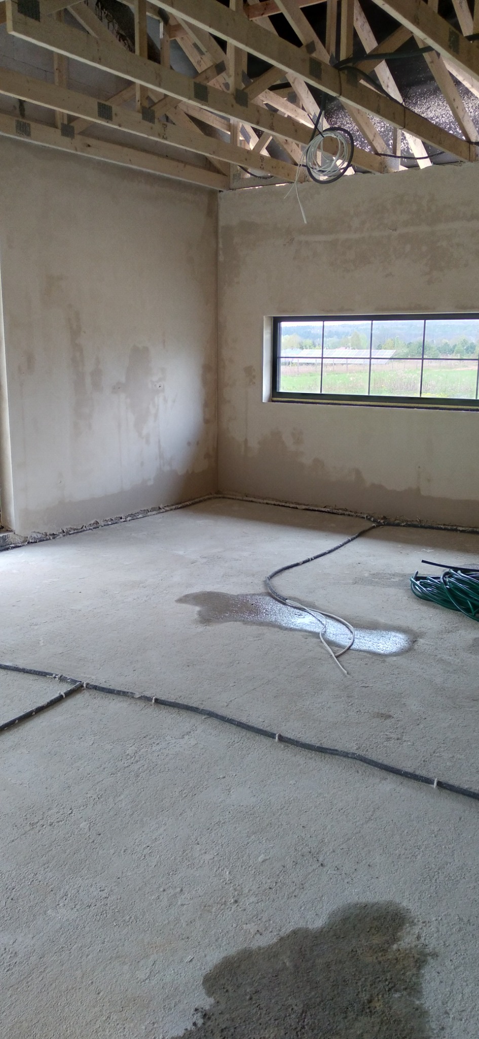 Surowe ściany i podłoga w trakcie prac wykończeniowych, widoczne kable na podłodze i drewniana konstrukcja dachu, okno z widokiem na zewnątrz.