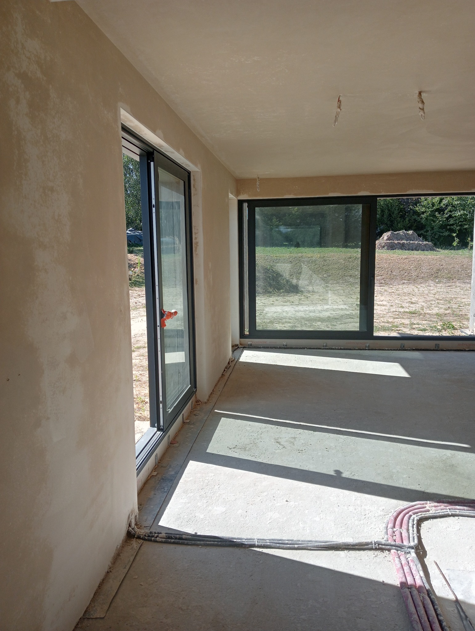 Wnętrze domu w trakcie tynkowania maszynowego ścian i sufitu, widoczne okna przesuwne z czarnymi ramami, wystające kable instalacji elektrycznej przy podłodze.