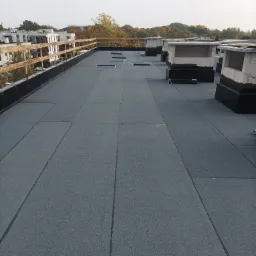 Płaski dach pokryty szarą papą termozgrzewalną, widoczne kominki wentylacyjne i drewniana balustrada na krawędzi dachu, w tle zabudowa miejska i drzewa.