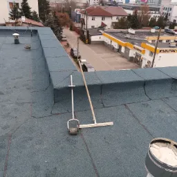 Widok z góry na płaski dach pokryty papą termozgrzewalną z widocznymi narzędziami do jej układania: wałek dociskowy i rozprowadzacz masy, w tle zabudowa miejska.